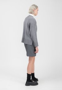 Grijze oversized trui met geribbelde mouwen, gecombineerd met een korte grijze rok met subtiele stippen. Zwarte plateau-schoenen en kniekousen.