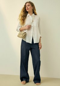 Mujer con largo cabello rubio lleva puesta una camisa blanca suelta abotonada, vaqueros anchos azul oscuro, bolso beige cruzado, sandalias y joyas de oro, de pie.