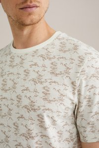 Svetle barevné tričko s jemnou texturou, zdobené hnědým abstraktním vzorem. Má klasický kulatý výstřih a krátké rukávy.