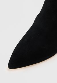Černé semišové kotníkové boty s špičatou špičkou a hladkou texturou. Podpatky jsou mírně nakloněné, což poskytuje elegantní siluetu.