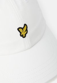 Căciulă albă dintr-un material texturat, cu un logo brodat al unei fluturi galben și negru pe partea din față. Detalii cusute sunt vizibile.