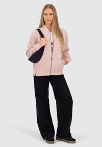 Giacca in pile zip con cappuccio rosa chiaro, borsa a tracolla nera, t-shirt grafica sotto, jeans larghi neri e scarpe nere con suole beige.
