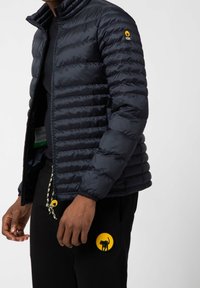 Granatowa kurtka puchowa z poziomym pikowaniem, zapięcie na zamek, wysoki kołnierz oraz wyraźna naszywka z logo na lewym rękawie.