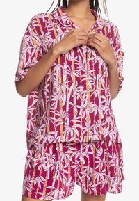 Krátký rukáv, košile na knoflíky a odpovídající šortky z růžové látky, zdobené bílými listy a žlutými akcenty v opakujícím se vzoru.