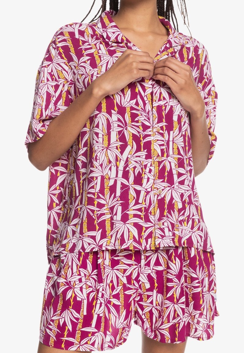 Krátký rukáv, košile na knoflíky a odpovídající šortky z růžové látky, zdobené bílými listy a žlutými akcenty v opakujícím se vzoru.