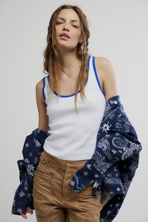 Jeune femme aux cheveux lâchés en tresse porte un débardeur blanc bordé de bleu, un pantalon marron et une veste bleue à motifs posée sur les épaules.