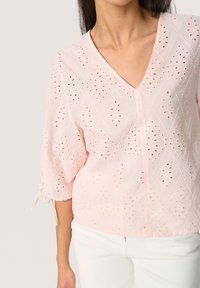 Blusa rosa chiaro con scollo a V, maniche a tre quarti, ricami a occhio in motivi a rombo e nastri alle estremità delle maniche.