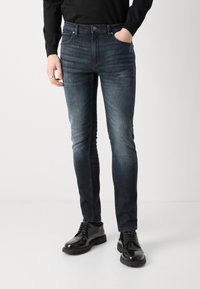 Jean skinny en denim foncé avec une finition délavée, doté de cinq poches, d'un bouton en métal, et porté avec des bottes noires à lacets.
