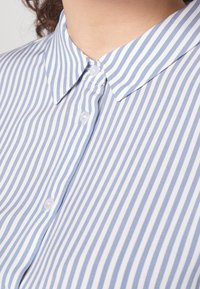 Camisa a rayas azul claro y blanca con cuello abotonado, que presenta puntas de cuello redondeadas y pequeños botones blancos a lo largo de la tapeta delantera.