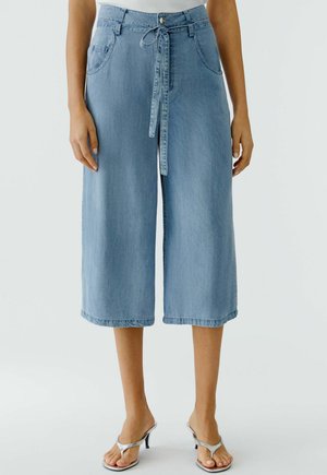 Vrouw draagt lichtblauwe denim culottes met wijde pijpen, voorzakken en een ceintuur, gecombineerd met zilverkleurige sandaalhakken.