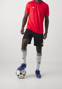 Röd idrottströja med gult Adidas-logotyp, svarta shorts, vita strumpor med svarta ränder och blå fotbollsskor på en svart och vit fotboll.