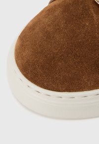 Καφέ καστόρινο παπούτσι με στρογγυλεμένη μύτη, λευκή λαστιχένια σόλα και εμφανή ραφές. Το άνω μέρος έχει μαλακή υφή και μινιμαλιστικό σχεδιασμό.