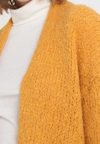 Knitted yellow cardigan with a textured finish, worn over a white ribbed turtleneck. Soft fabric and open-front design.