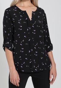 Černá blůza s výstřihem do V a ohrnutými rukávy, s květinovým vzorem v purpurové a bílé. Lehátkový materiál, volný střih, detail s knoflíkem.