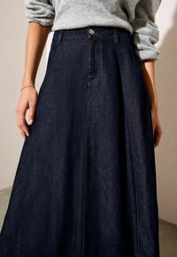 Sötétkék farmer szoknya magas derékkal, gombos záródással, redőzéssel és sima textúrával. Világosszürke pulóverrel összeállítva.
