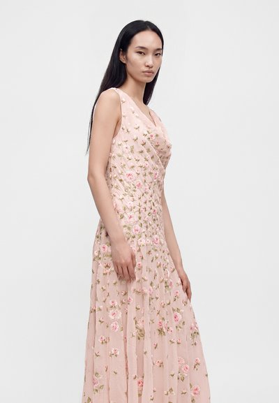 Femme aux longs cheveux noirs portant une robe rose pâle sans manches avec broderies florales, debout devant un fond clair uni.