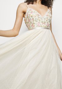 Šaty s květinovou výšivkou, V-krkem a ramínky typu spaghetti, s průsvitným puntíkovaným přehozem v krémové barvě a saténovým pasem.