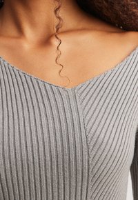Haut en tricot gris côtelé avec un décolleté en V, présentant une finition texturée et de subtils détails de couture sur l'avant, mettant en valeur sa silhouette ajustée.