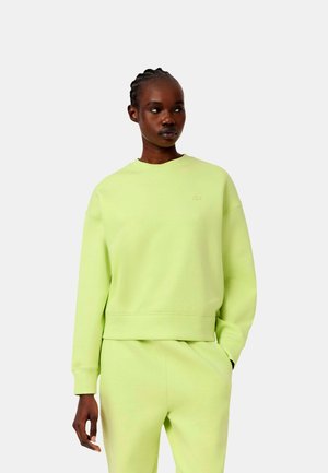 Mujer con el cabello trenzado que lleva una sudadera verde claro y pantalones a juego, de pie con una mano en el bolsillo, mirando hacia un lado.