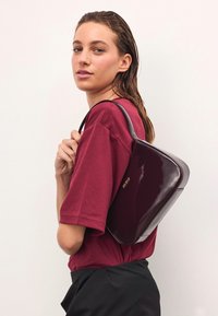 Spīdīga burgundijas krāsas rokassoma ar strukturētu formu, ar elegantu melnu siksnu un diskretu zelta zīmolu, turēta pretī bordo kreklam.