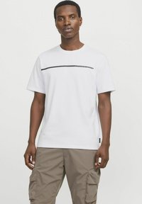 Hvit bomull T-skjorte med korte ermer, med en horisontal svart stripe og branding. Brukt med lysebrune cargo-bukser.