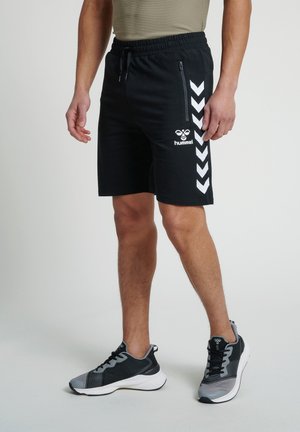 Man in zwarte hummel sportshorts met witte chevronstrepen en zwart-grijze sportschoenen, staand op een effen achtergrond.