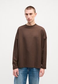 Chemise marron à manches longues en coton, avec un col rond et une coupe décontractée. Logo discret sur la poitrine, associée à un jean bleu.