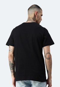Schwarzes Baumwoll-T-Shirt mit kurzen Ärmeln, rundem Halsausschnitt und glatter Textur. Sichtbare Tätowierungen an den Armen, keine auffälligen Designelemente.