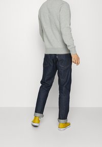 Sudadera gris, vaqueros de mezclilla azul oscuro y zapatos de lona amarillos. Los vaqueros están remangados en los puños, mostrando los detalles de los zapatos.