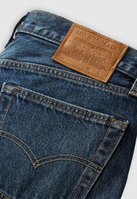 Blue denim jeans featuring a leather branded patch at the waistband, contrasting yellow stitching, and curved rear pocket design.