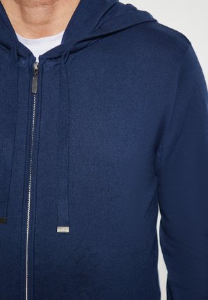 Marineblaue Zip-Hoodie aus strukturiertem Stoff mit Kapuze und Kordelzug, ausgestattet mit silbernen Beschlägen und einem durchgehenden Reißverschluss.