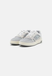 Sneakers grigi e bianchi con tessuto a rete, suola in gomma solida, design stringato e dettagli del logo in evidenza sul lato. Superfici lisce e testurizzate.