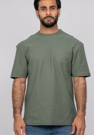 Homme barbu portant un t-shirt manches courtes vert olive avec une poche poitrine et un jean bleu, debout devant un fond gris clair.