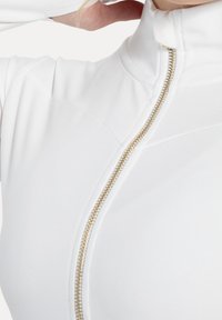 White zip-up top featuring a high collar, gold zipper, and smooth fabric texture. Close-up view highlights the zipper detail and design lines.