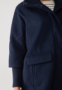 Marineblauer Wollmischmantel mit einem Reißverschlusshals, seitlichen Taschen und gerippten Bündchen. Weiche Textur und strukturiertes Design.