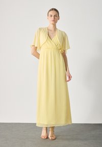 Light yellow maxi dress with flutter sleeves and a deep V-neck, featuring a cinched waist and flowing skirt. Model standing on a gray surface.