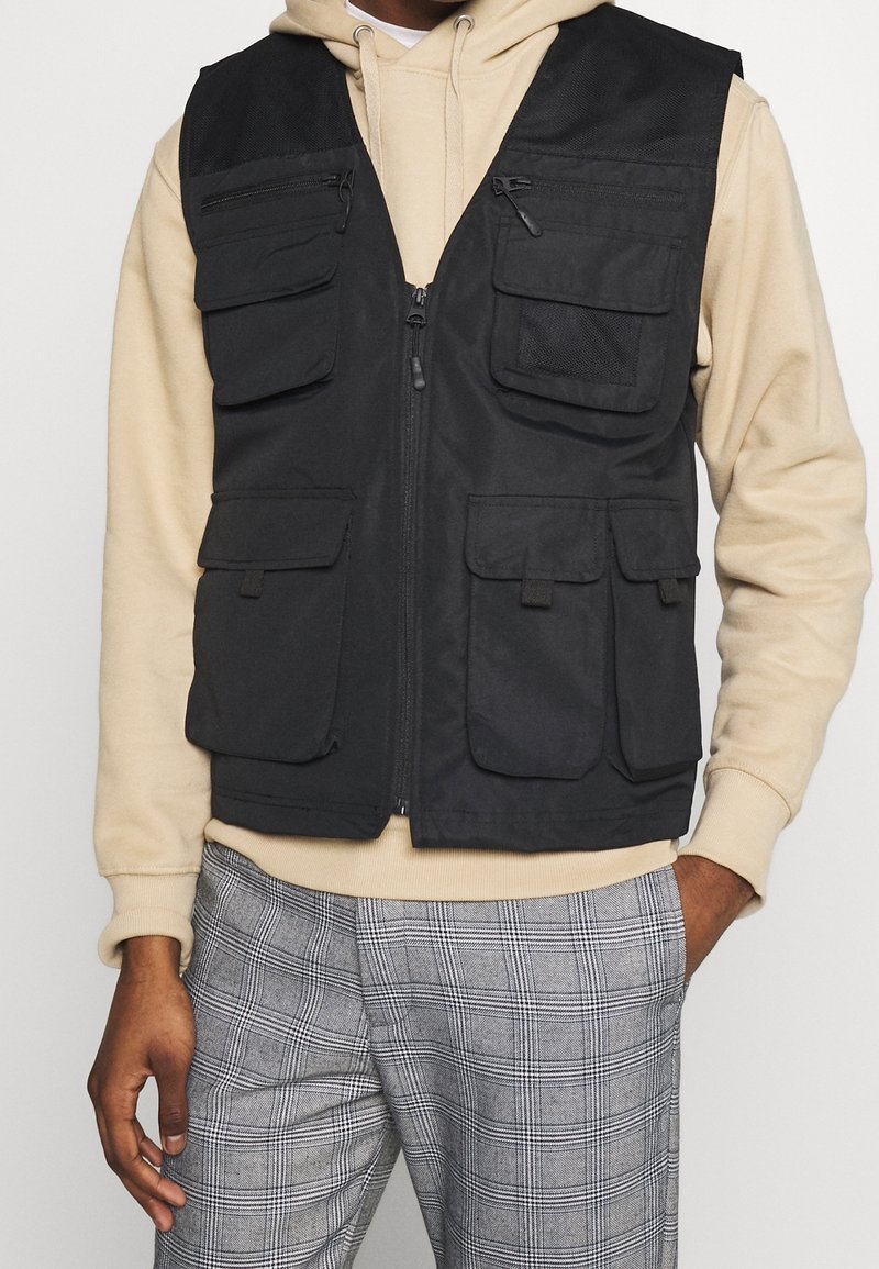Persona con sudadera beige, chaleco negro de múltiples bolsillos con cremallera y pantalones de cuadros grises, de pie con una mano en el bolsillo.