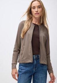 Ljusbrun zip-up jacka med en slät textur, sidofickor och rund halsringning, matchad med blå jeans och en mörkbrun topp.