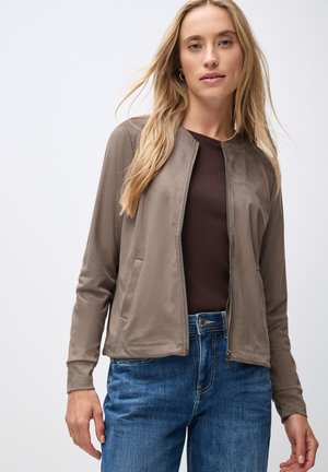 Light brown zip-up jacket with a smooth texture, side pockets, and a round neckline, paired with blue jeans and a dark brown top.