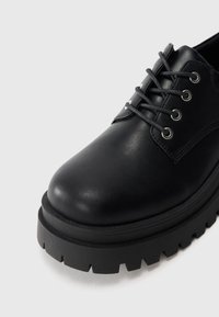 Chaussure noire en cuir à lacets avec semelle épaisse et volumineuse et œillets en métal, vue de près sous un angle avant.