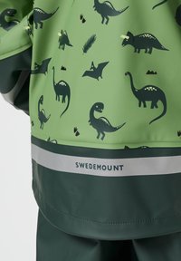 Enfant portant un imperméable vert avec des motifs de dinosaures foncés, une bande réfléchissante et le texte SWEDEMOUNT près de l’ourlet.