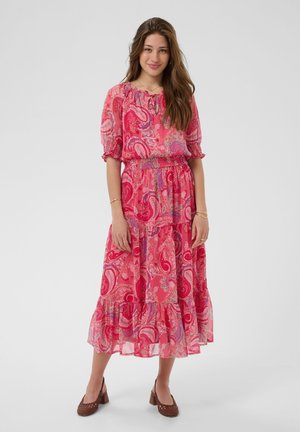 Giovane donna con capelli lunghi che indossa un vestito midi rosa con motivo paisley e scarpe marroni con tacco a blocco, in piedi davanti a uno sfondo semplice.