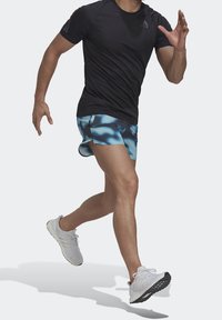 T-shirt de sport noir à manches courtes, shorts à motifs bleus et chaussures de course blanches. La silhouette est en position de course sur un fond uni.