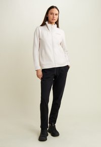 Donna in piedi che indossa una felpa bianca con zip e pantaloni sportivi neri con scarpe da ginnastica nere su uno sfondo semplice.