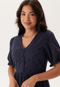 Abito blu navy con scollatura a V, maniche corte a sbuffo, motivo floreale a intaglio e dettagli con bottoni lungo la parte anteriore. Tessuto morbido e design fluido.