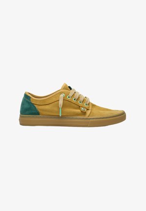 Zapatilla casual de corte bajo con cuerpo de ante color mostaza, talón de ante verde, cordones beige y suela de goma marrón claro.
