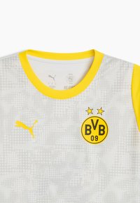 Camiseta blanca con patrón de puntos grises, cuello amarillo y acentos en las mangas. Presenta el logo amarillo de Puma y el escudo del Borussia Dortmund con estrellas.