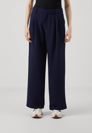 Navy wide-leg trousers with pleats, made of soft fabric. Features a high waistband and pockets, paired with light-colored sneakers.