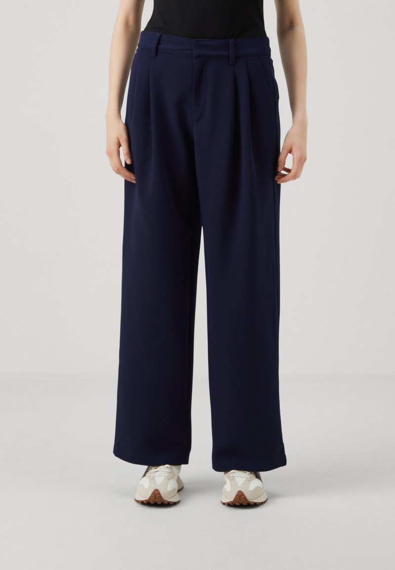 Navy wide-leg trousers with pleats, made of soft fabric. Features a high waistband and pockets, paired with light-colored sneakers.