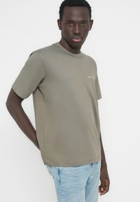 Λαδί βαμβακερό μπλουζάκι με στρογγυλή λαιμόκοψη, κοντά μανίκια και διακριτικό λογότυπο στο στήθος, συνδυασμένο με ανοιχτό μπλε τζιν παντελόνι.